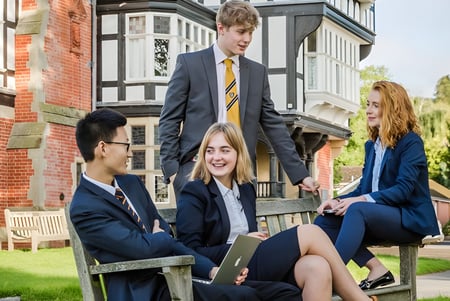 Eine Gruppe von Schülern sitzt auf einer Bank im Freien auf dem Gelände des Bedstone College vor einem Backsteingebäude.
