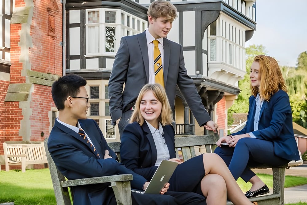 Eine Gruppe von Schülern sitzt auf einer Bank im Freien auf dem Gelände des Bedstone College vor einem Backsteingebäude.