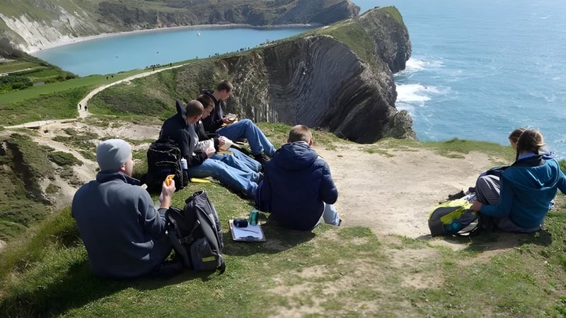Schüler der Beech Grove School wandern auf einem grasbewachsenen Pfad mit Blick auf eine felsige Küstenlandschaft und das türkisfarbene Meer.