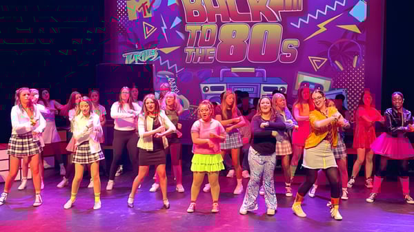 Eine Gruppe Schülerinnen des Beech Hill College steht in bunten Outfits auf der Bühne vor einem retro 'Back to the 80s' Hintergrund.