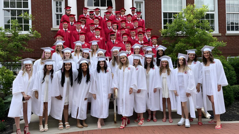Eine große Gruppe von Schülerinnen und Schülern der Beechwood Sacred Heart School steht in roten und weißen Talaren vor einem Backsteingebäude.