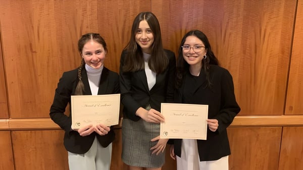 Drei Frauen in formeller Kleidung halten Zertifikate vor einer Holzvertäfelung auf dem Campus der Bellarmine Preparatory School.