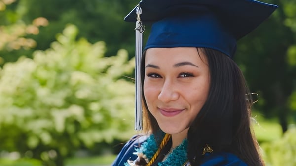 Eine Absolventin der Bellarmine Preparatory School in blauer Abschlusskleidung steht in einer grünen Außenanlage und lächelt.