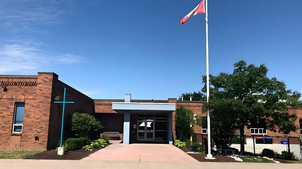 Das Backsteingebäude der Belle River District High School mit einer großen kanadischen Flagge über dem Eingang und Bäumen im Vordergrund.