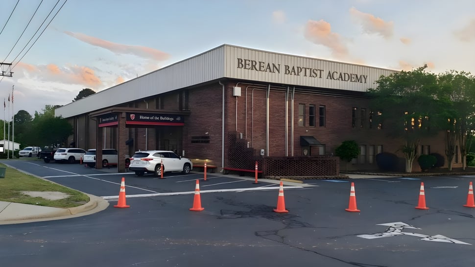 Das Backsteingebäude der Berean Baptist Academy ist von einem Parkplatz mit Fahrzeugen und Absperrungen umgeben.