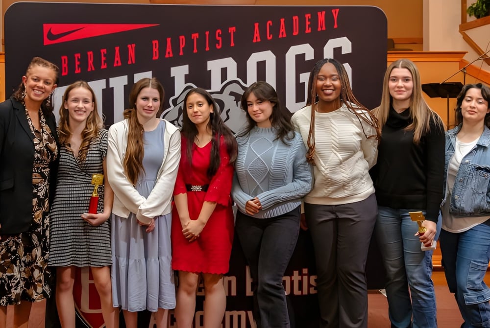 Eine Gruppe Schülerinnen steht vor einem Banner der Berean Baptist Academy in verschiedenen Kleidungsstilen.