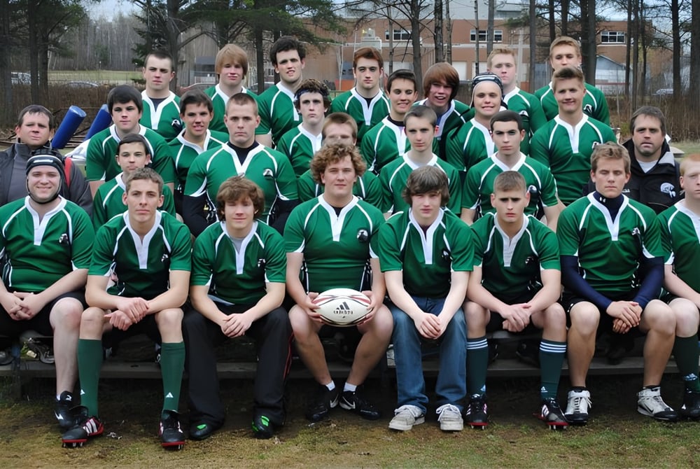 Eine Gruppe von Schülerinnen und Schülern der Bernice McNaughton High School sitzt in Sportuniformen auf einem Feld vor einem Wald.