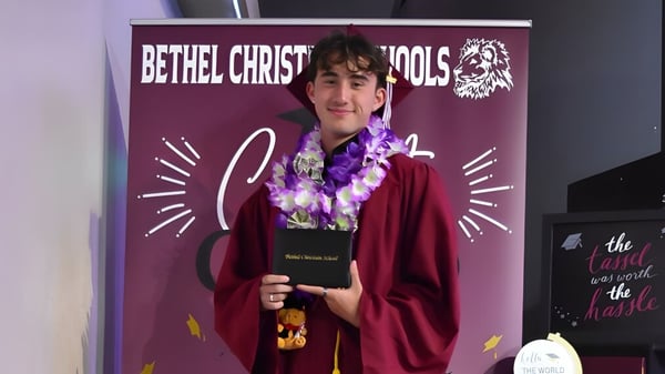 Eine Person in rotem Gewand mit Blumenkranz steht vor einem lila Banner der Bethel Christian Schools mit Löwenlogo.