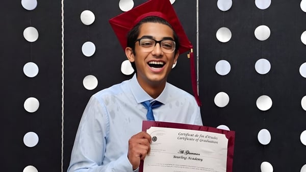 Ein Absolvent der Beurling Academy hält in seinem Abschlussmantel und -hut ein Diplom vor einem schwarz-weißen gepunkteten Hintergrund.