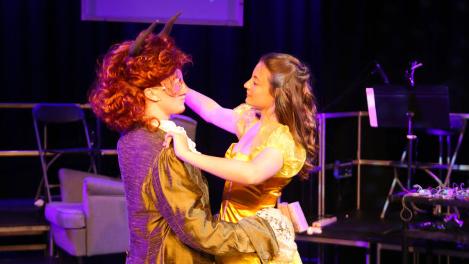 Zwei Frauen in bunten Kleidern umarmen sich auf der Bühne des Bexhill College vor lila Hintergrund.