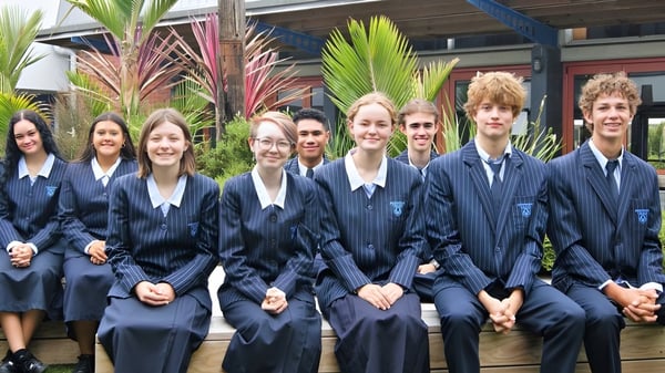 Eine Gruppe von Schülerinnen und Schülern in Schuluniform steht vor dem Gebäude des Birkenhead College mit tropischen Pflanzen im Hintergrund.