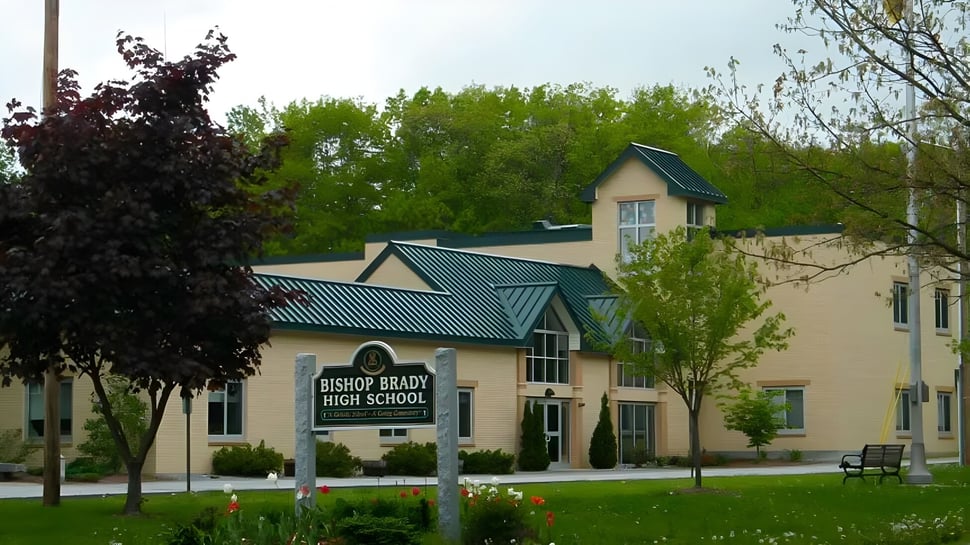 Das zweistöckige Backsteingebäude mit grünem Metalldach gehört zur Bishop Brady High School und ist von Bäumen und Wiese umgeben.