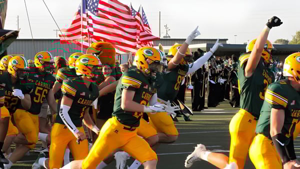 Eine Gruppe Footballspieler der Bishop Carroll High School steht mit amerikanischen Flaggen auf dem Spielfeld vor einem Stadion.