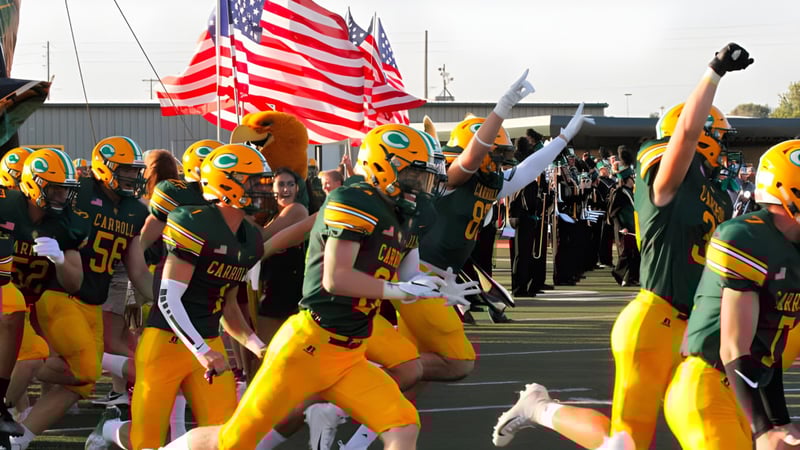 Eine Gruppe Footballspieler der Bishop Carroll High School steht mit amerikanischen Flaggen auf dem Spielfeld vor einem Stadion.