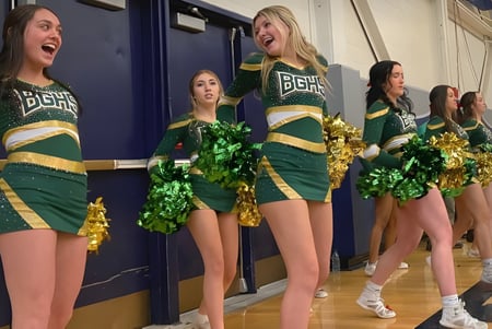Eine Gruppe Cheerleader in grünen und goldenen Uniformen steht mit Pompons im Flur der Bishop Guertin High School.