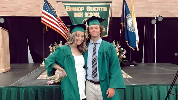 Zwei Schüler der Bishop Guertin High School stehen in Abschlusskleidung vor einer Kulisse mit amerikanischer und Schulflagge.