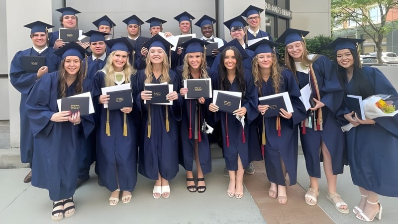 Eine Gruppe von Absolventen der Bishop Heelan Catholic High School steht im Freien in akademischer Kleidung und hält ihre Zeugnisse.