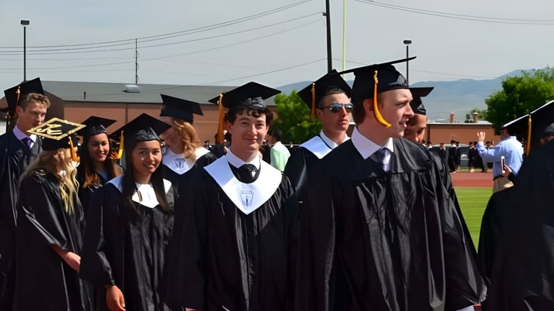 Eine Gruppe von Absolventinnen und Absolventen der Bishop Kelly High School steht im schwarzen Abschlussgewand auf einem Feld vor einem Gebäude und Bergen.