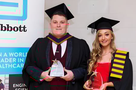 Zwei Absolventen in Talaren stehen vor einem Abbott-Logo auf dem Campus der Blackwater Community School.