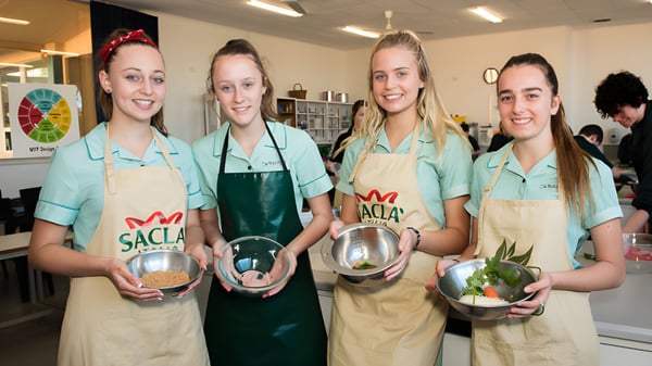 Vier Schülerinnen der Blackwood High School tragen grüne Schürzen und halten Kochutensilien in einer Küche.