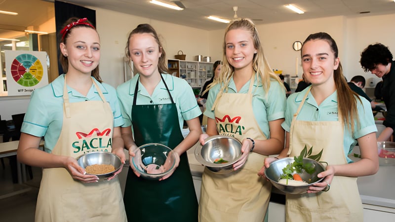 Vier Schülerinnen der Blackwood High School tragen grüne Schürzen und halten Kochutensilien in einer Küche.