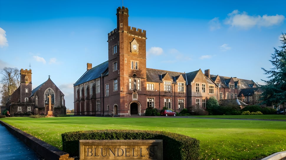 Das historische Backsteingebäude mit hohem Uhrenturm der Blundells School steht vor gepflegtem Rasen und grüner Umgebung.