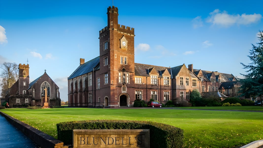 Das historische Backsteingebäude mit hohem Uhrenturm der Blundells School steht vor gepflegtem Rasen und grüner Umgebung.