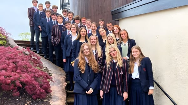 Eine große Gruppe von Schülerinnen und Schülern der Blundells School steht in formeller Kleidung vor einem Holzgebäude mit blühenden Blumen.