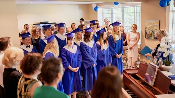 Eine Gruppe Absolventen der Blyth Academy steht in blauen Talaren und Hüten zusammen in einem Raum mit einem Klavier und Blumendekoration.