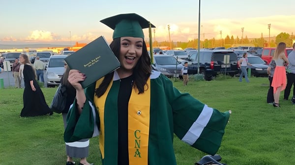 Eine Absolventin der Bonneville High School trägt grüne Abschlusskleidung und hält ein Diplom vor einem Parkplatz.