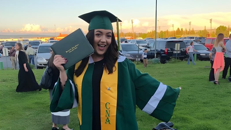 Eine Absolventin der Bonneville High School trägt grüne Abschlusskleidung und hält ein Diplom vor einem Parkplatz.