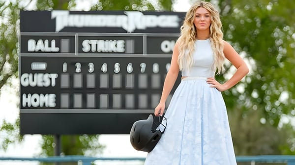 Eine junge Frau steht vor der Anzeigetafel eines Baseballspiels auf dem Gelände des Bonneville Joint School District und hält einen schwarzen Hut.