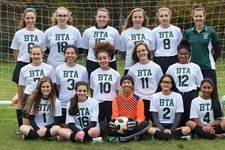 Eine Gruppe junger Fußballspielerinnen der Boston Trinity Academy posiert gemeinsam auf einem Grasfeld mit einem Fußball im Vordergrund.