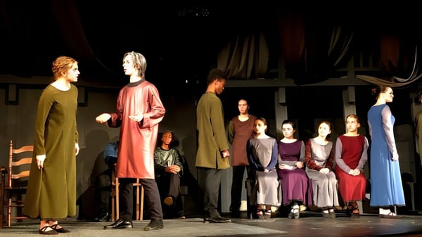 Schüler der Boston Trinity Academy stehen in bunten Kostümen auf einer Bühne mit Theaterkulisse.