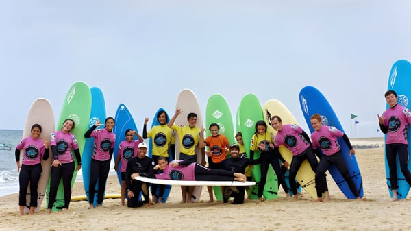 Schüler des Bosworth Independent College stehen mit bunten Neoprenanzügen und Surfbrettern am Strand vor dem Ozean.
