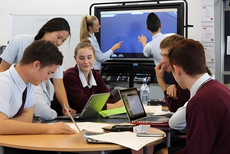 Eine Gruppe von Schülerinnen und Schülern der Botany Downs Secondary College diskutiert und arbeitet mit elektronischen Geräten an einem Tisch.