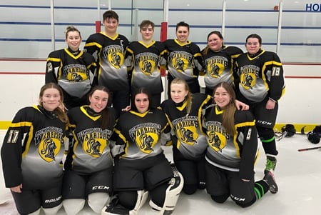 Eine Gruppe von jungen Hockeyspielerinnen der Botwood Collegiate posiert gemeinsam auf der Eisbahn mit dem Schriftzug HOME im Hintergrund.