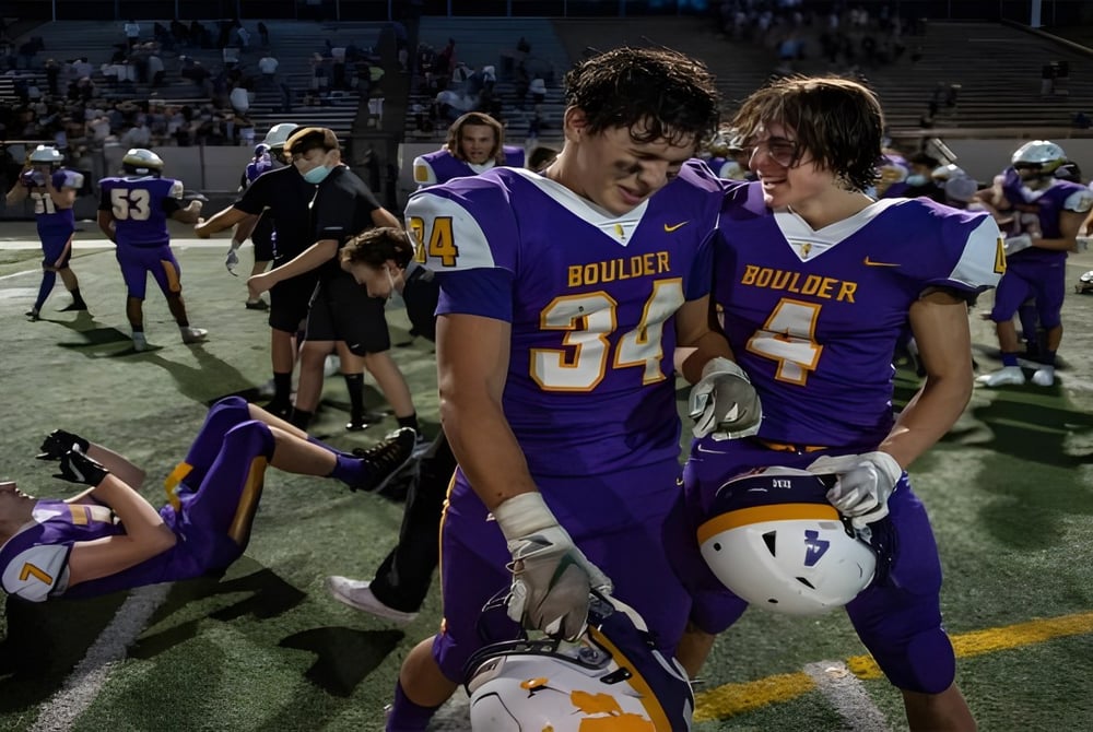 Zwei Footballspieler stehen auf dem Spielfeld der Boulder Valley Public Schools mit Spielern und Zuschauern im Hintergrund.