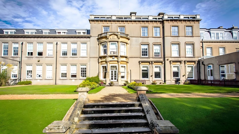 Das Hauptgebäude der Bournemouth Collegiate School mit gepflegtem Rasen und steinernen Treppen unter blauem Himmel.