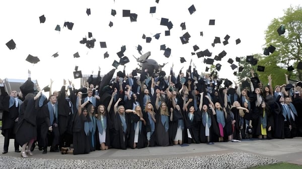 Eine Gruppe von Absolventen in schwarzen Roben wirft ihre Hüte auf dem Campus des Bradfield College in die Luft vor grünem Hintergrund.