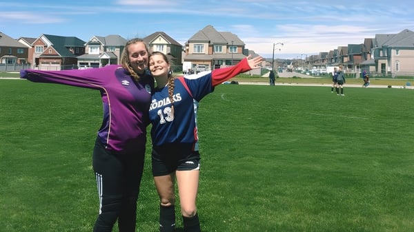 Zwei Schülerinnen der Bradford District High School umarmen sich auf einem Sportplatz vor bunten Häusern.