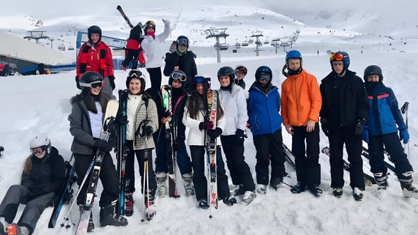 Eine Gruppe von Schülerinnen und Schülern des Braemar College steht in Winterkleidung mit Skiausrüstung auf einem verschneiten Hang vor Bergen und Skiliften.