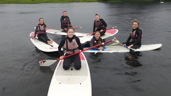 Schüler des Breifne College stehen in Neoprenanzügen auf Paddleboards im Wasser umgeben von grüner Natur.