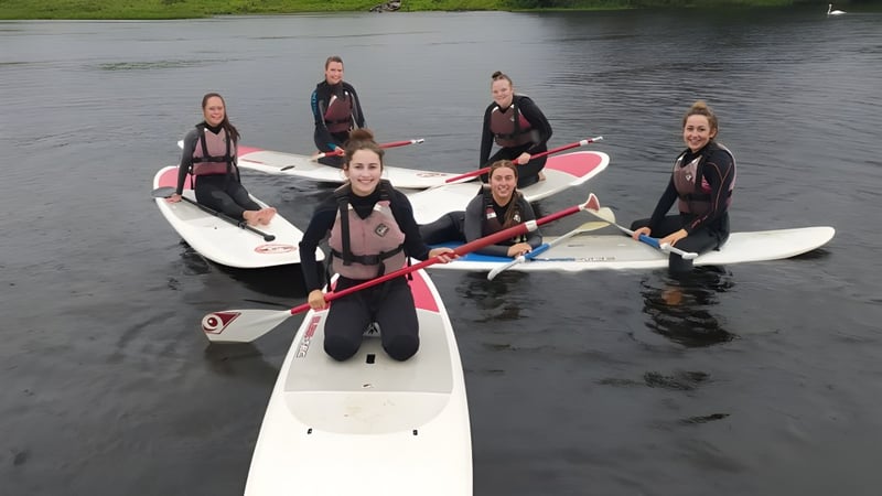 Schüler des Breifne College stehen in Neoprenanzügen auf Paddleboards im Wasser umgeben von grüner Natur.