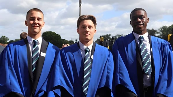 Drei Schüler der Brentwood School in blauen Abschlussroben stehen vor bewölktem Himmel mit Bäumen im Hintergrund.