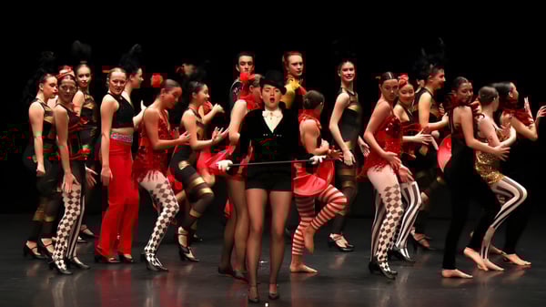 Schüler des Brighton College treten in farbenfrohen Kostümen auf einer dunklen Bühne auf.