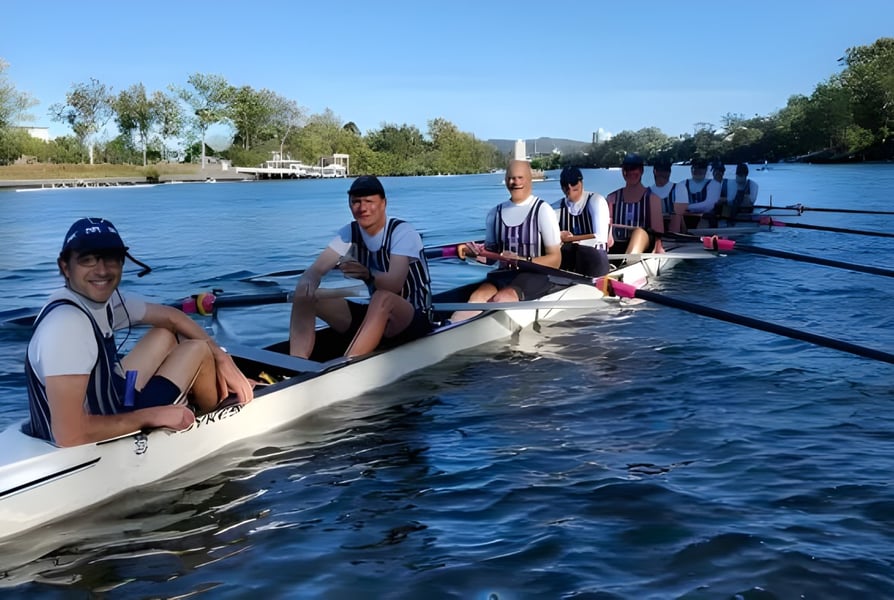 Eine Gruppe von Schülerinnen und Schülern der Brisbane Grammar School beim Rudern auf einem Fluss, umgeben von Bäumen unter blauem Himmel.