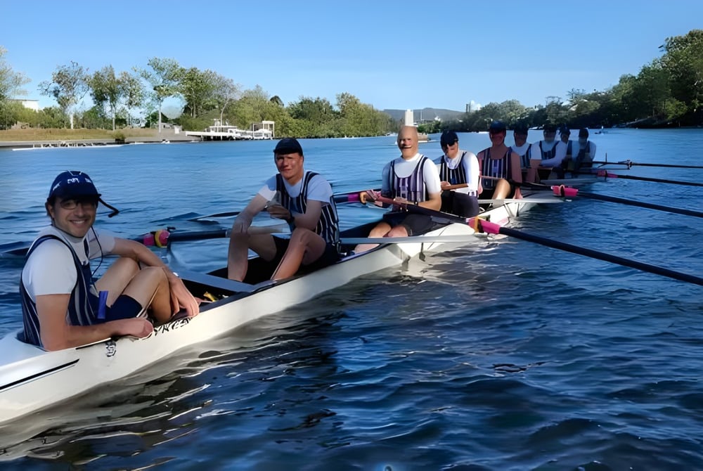 Eine Gruppe von Schülerinnen und Schülern der Brisbane Grammar School rudert auf einem Fluss, umgeben von Bäumen und blauem Himmel.