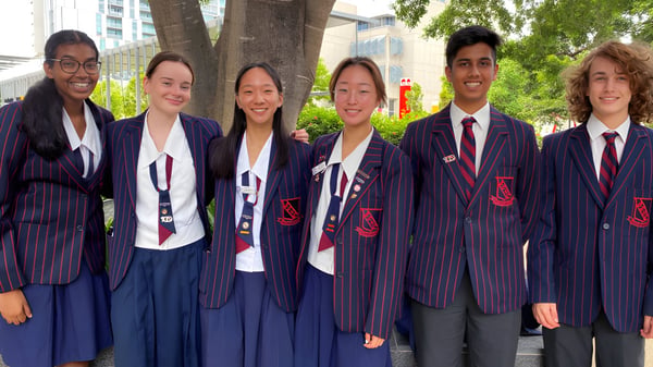 Eine Gruppe Schüler der Brisbane State High School steht vor einem baumbestandenen städtischen Hintergrund.