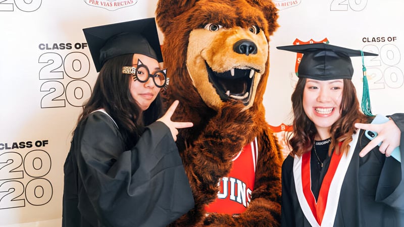 Zwei Schülerinnen der Britannia Secondary School in Abschlussroben posieren mit dem Maskottchen der Schule vor dem Class of 2020 Hintergrund.